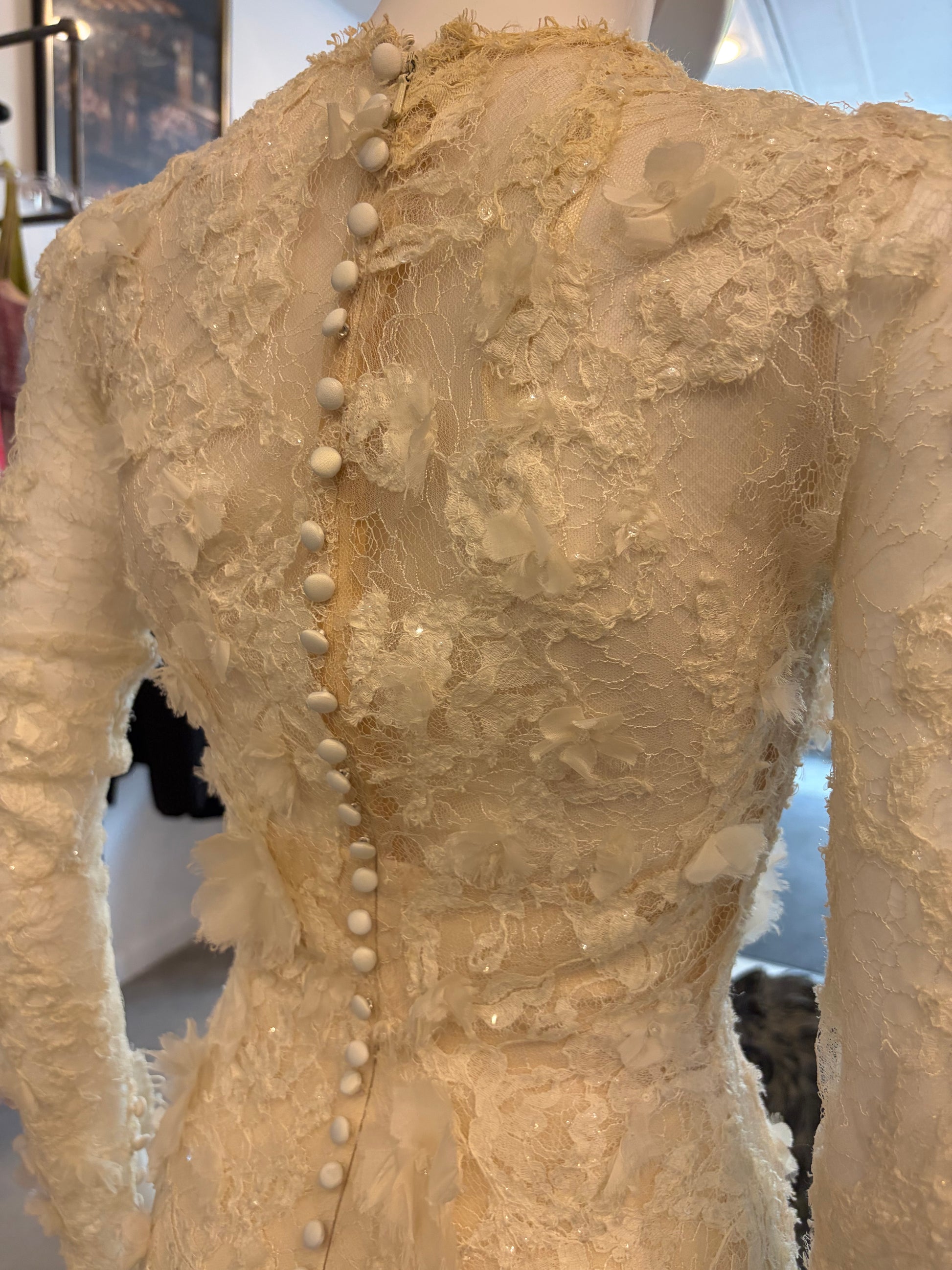Close-up back view of a cream-colored lace dress on a mannequin, featuring textured floral lace, long sleeves, and a vertical row of decorative pearl-like buttons down the center.